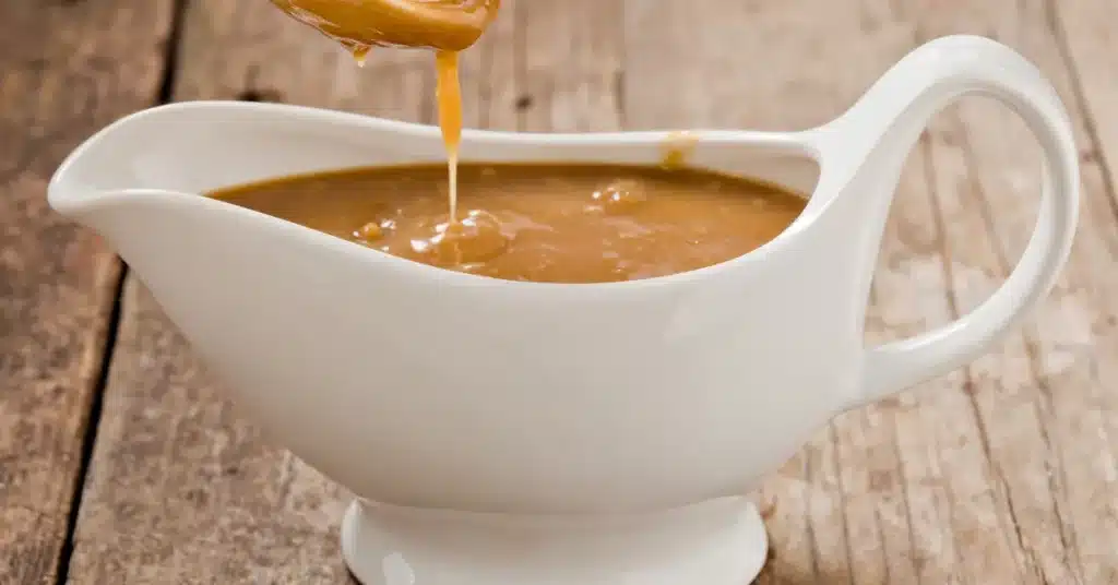A close up shot of a small white ladle pouring brown gravy into a white ceramic gravy boat. Shot on a grungy old wooden table.