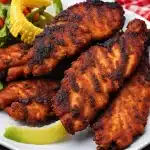 A white plate topped with blackened chicken tenders and corn on the cob on a table.
